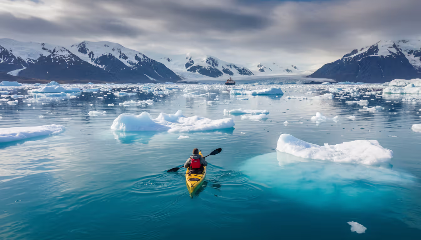 Solo kayaker in safety gear paddling among icebergs with a small expedition ship and snowy mountains in the distance