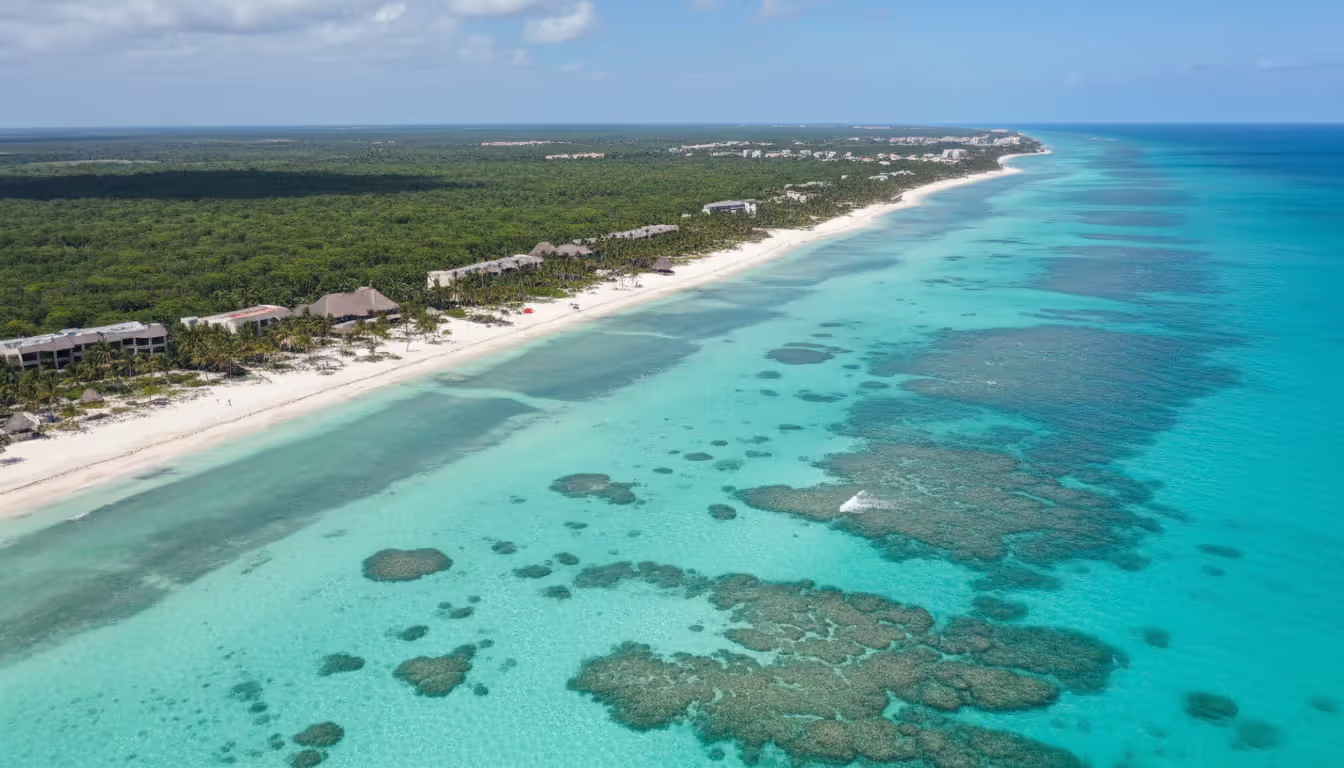 Aerial view of Riviera Maya coastline with turquoise Caribbean water, coral reef visible through clear sea, white sand beach, palm trees, and resort buildings nestled in tropical jungle