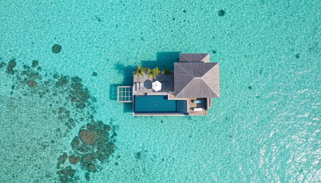 Aerial view of a luxury overwater villa with private infinity pool and glass floor panels surrounded by clear turquoise ocean water and coral reefs