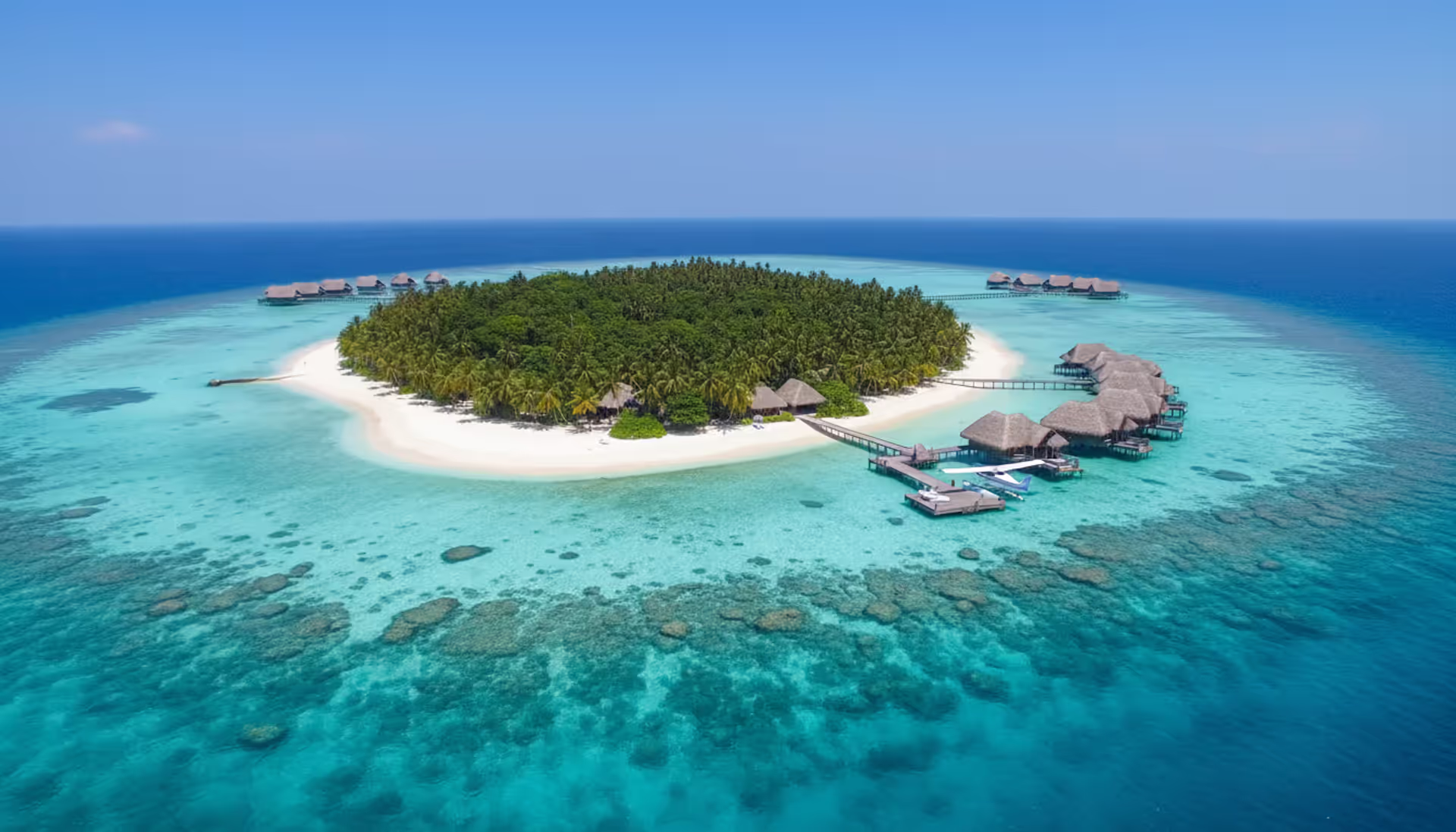 Aerial view of a tropical private island with turquoise lagoon, overwater luxury villas, white sandy beach, and a seaplane docked at a wooden pier on a sunny day